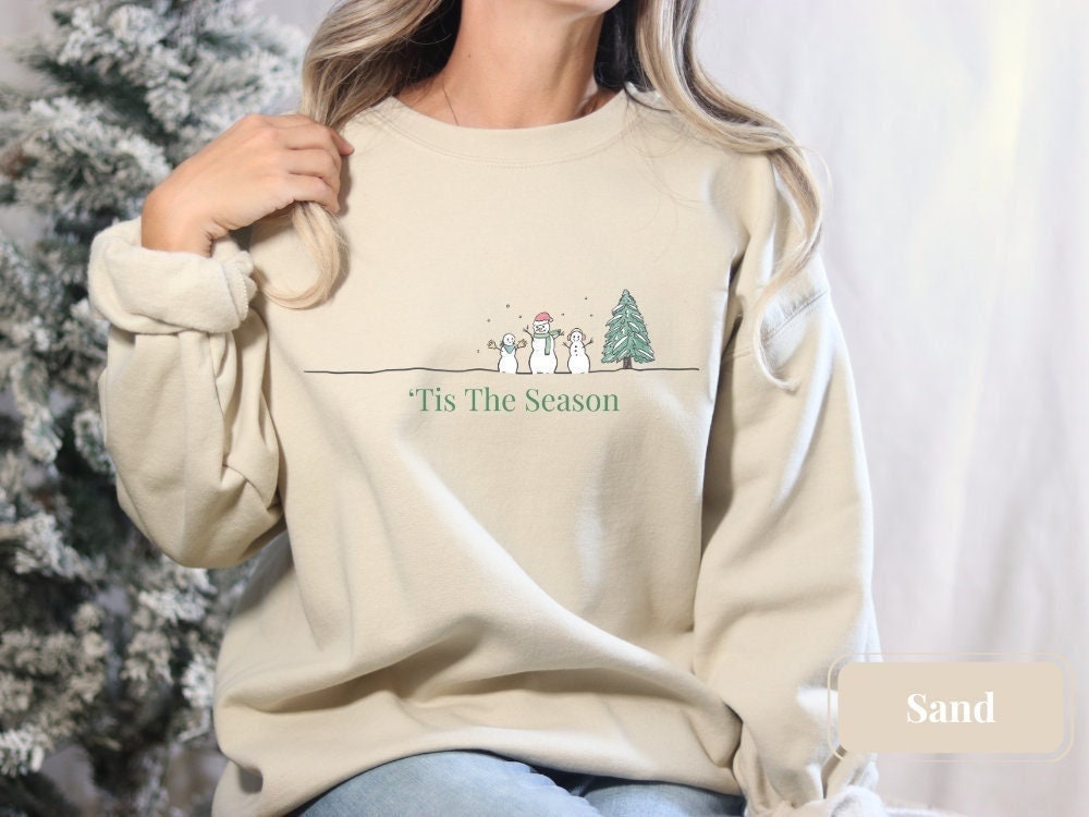 Minimalist sand Christmas crewneck sweatshirt on a model, adorned with a simple snowman and Christmas tree design, captioned 'Tis The Season.' Perfect for holiday festivities or as a cozy winter sweatshirt.