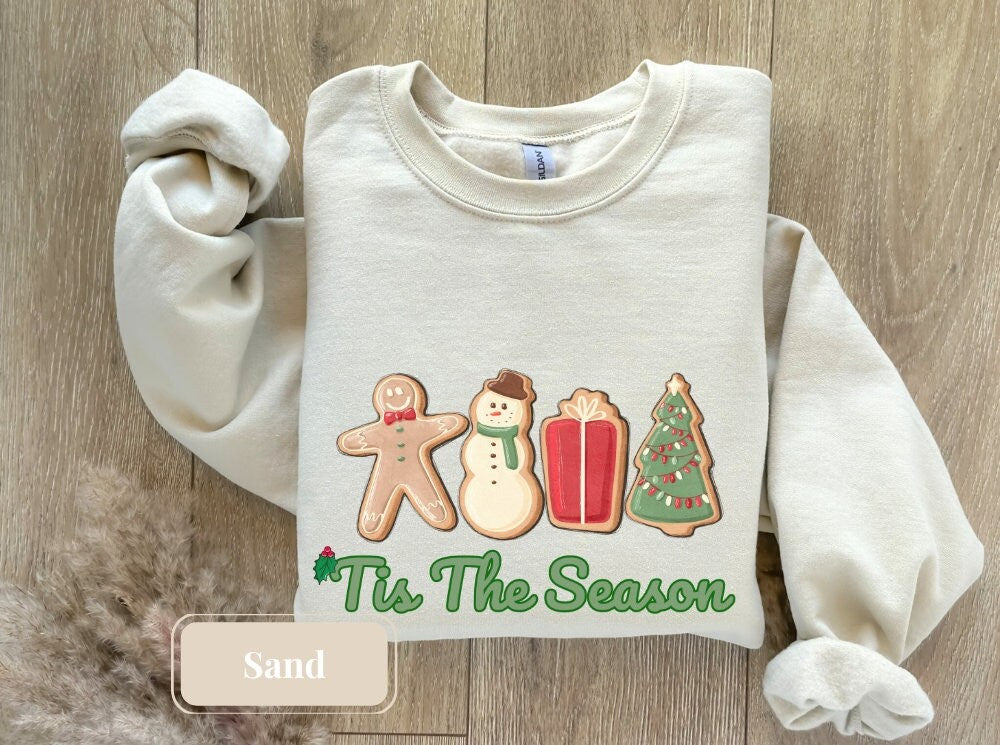 Sand-colored Christmas sweatshirt showcasing gingerbread man, snowman, present, and Christmas tree with 'Tis The Season' in green, a must-have for cookie lovers and those celebrating the festive, holiday spirit.