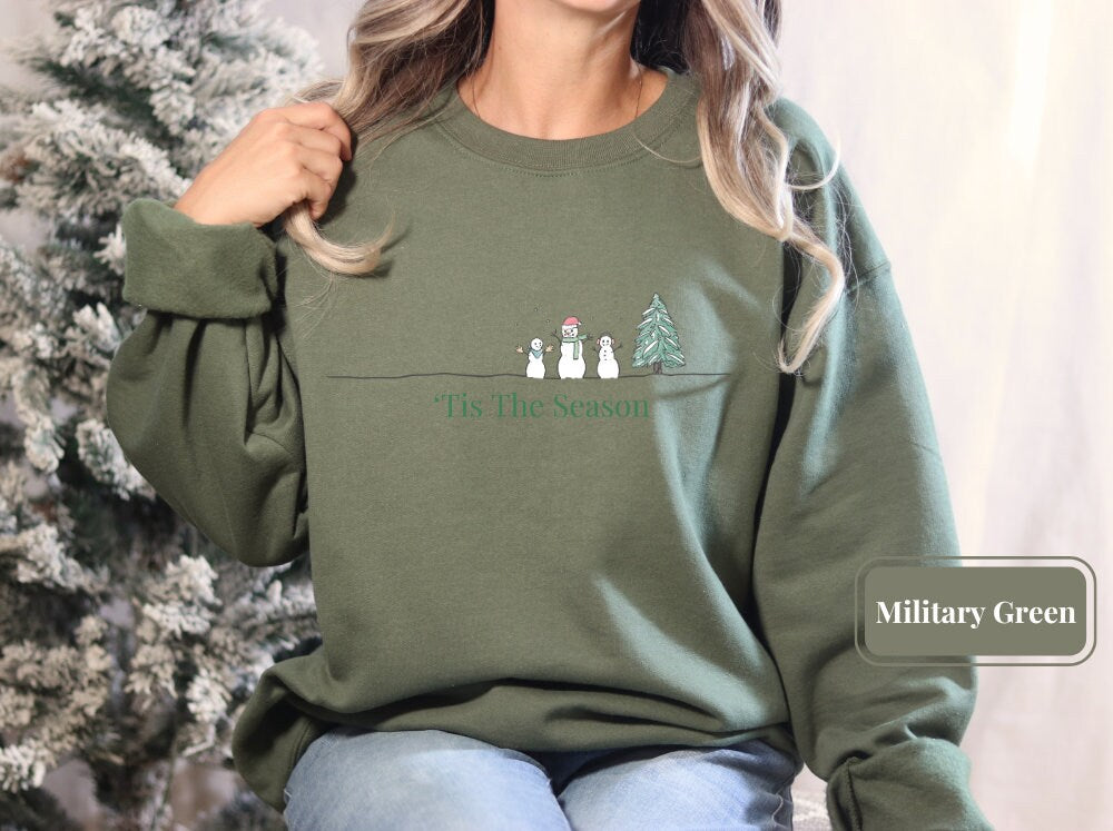 Military green holiday sweatshirt with a festive snowman and Christmas tree design, captioned 'Tis The Season.' Perfect for casual Christmas gatherings, offering both comfort and a touch of holiday spirit.