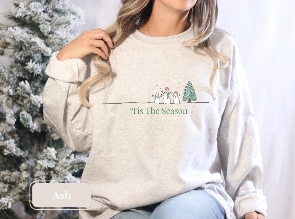 Cozy ash-colored Christmas sweatshirt featuring a minimalist design with a snowman, tree, and 'Tis The Season' text. Ideal for holiday parties, comfy and stylish, perfect as a Christmas gift or winter outfit.