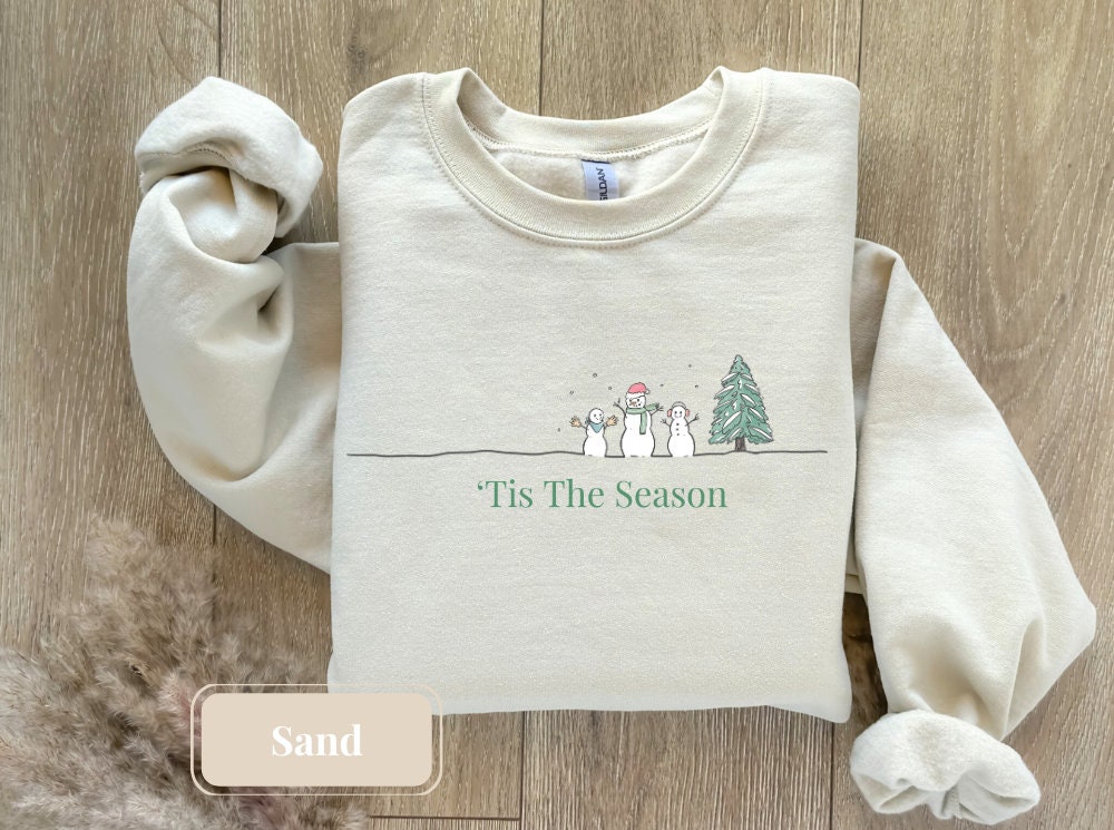 Soft sand-hued crewneck displaying a charming Christmas scene with a snowman and tree, understated 'Tis The Season' text. A minimalist Christmas sweater ideal for gifting or seasonal wear.