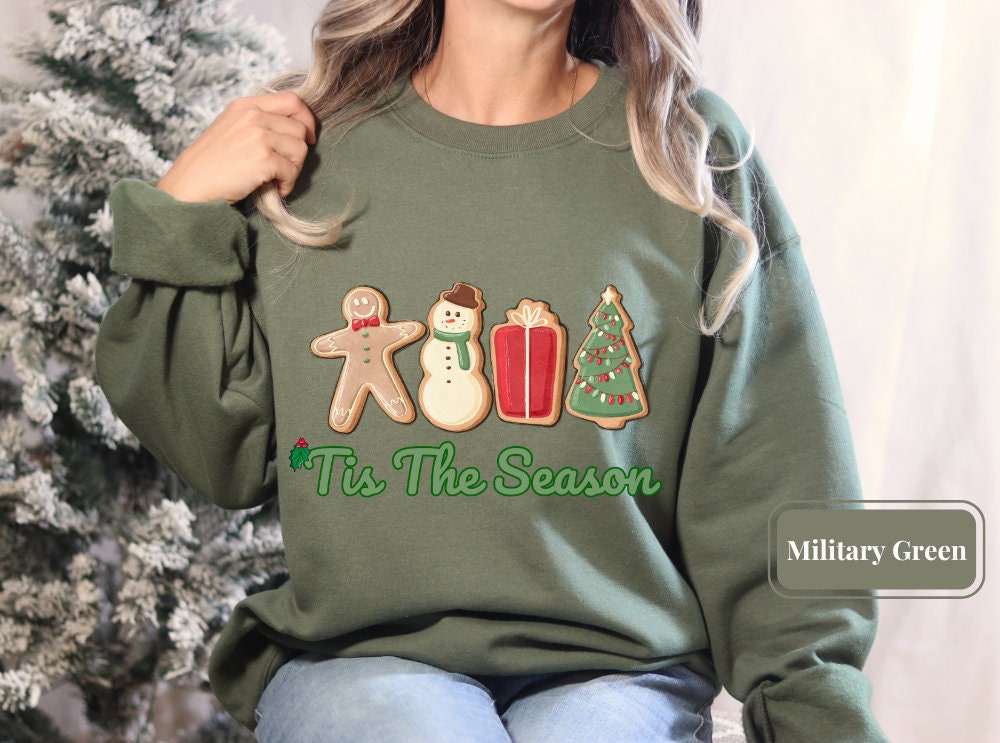 Military green Christmas sweatshirt adorned with colorful gingerbread, snowman, gift, and tree, captioned 'Tis The Season' for those celebrating peace, love, and the New Year, ideal for Christmas squad members.