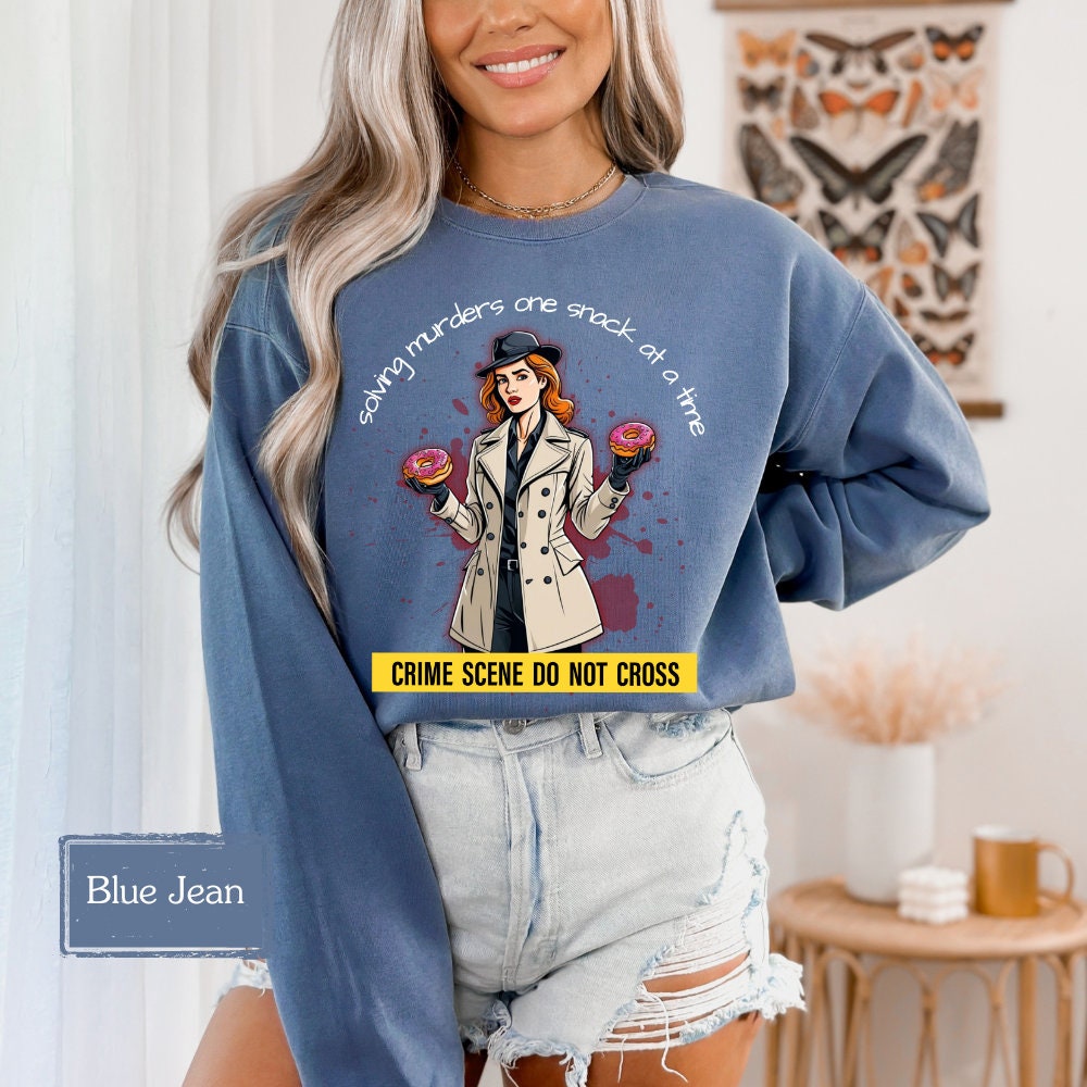 Blue Jean-colored sweatshirt with a funny murder mystery design, featuring a detective with donuts and the phrase &#39;solving murders one snack at a time.&#39; A cozy and stylish choice for crime show fans and snack lovers alike.