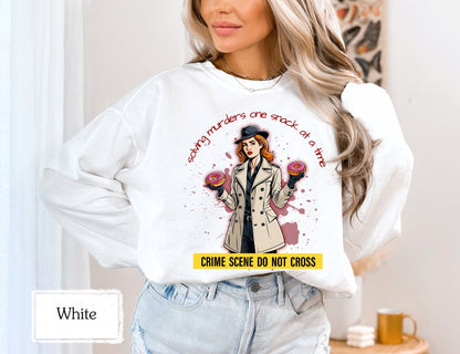 White sweatshirt with a crime-themed design, showing a detective holding donuts and the words &#39;solving murders one snack at a time.&#39; Perfect for true crime enthusiasts and crime junkies who love humor and comfort.