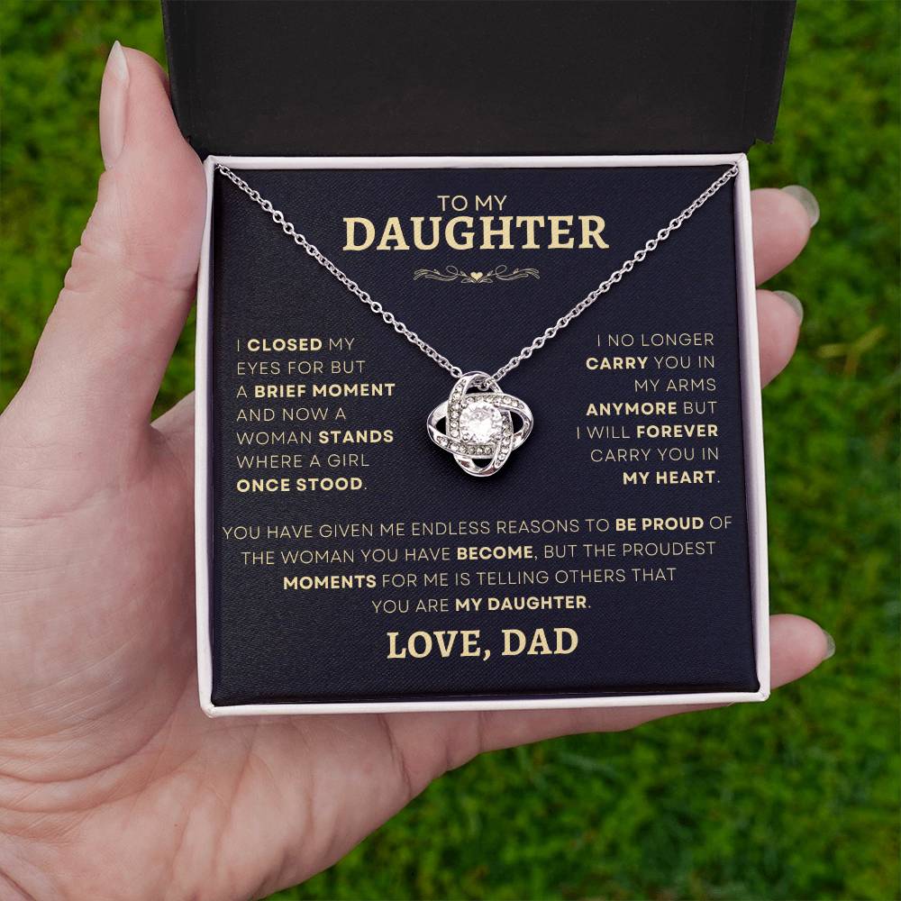 Hand holding an open gift box with 'To My Daughter' Silver Love Knot Necklace on a grassy background, showcasing a touching message from dad, perfect for expressing lifelong fatherly love and pride.